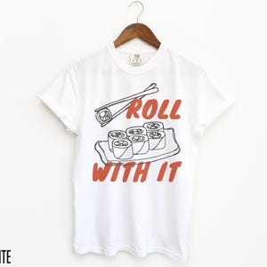 May include: White t-shirt featuring a graphic of sushi rolls and chopsticks. The text "Roll with it" is printed in red on the shirt.