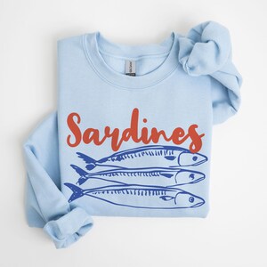 May include: A light blue sweatshirt with the word "Sardines" printed in red on the front. Three blue sardines are pictured below the word.