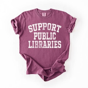 May include: A maroon t-shirt with rolled-up sleeves. The shirt features the text "SUPPORT PUBLIC LIBRARIES" in a distressed, white, collegiate-style font. The shirt is displayed on a white background.