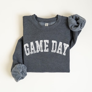 May include: A dark gray sweatshirt with the text "GAME DAY" in distressed white lettering across the chest. The sleeves are cuffed, and the sweatshirt is laid flat on a white surface. The sweatshirt is a crew neck style.