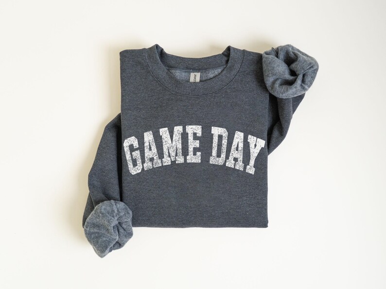 May include: A dark gray sweatshirt with the text "GAME DAY" in distressed white lettering across the chest. The sleeves are cuffed, and the sweatshirt is laid flat on a white surface. The sweatshirt is a crew neck style.