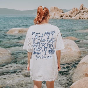 May include: White t-shirt with a blue graphic design that says "Miami Beach + Girl's Trip 2025" and includes images of drinks, sunglasses, flip flops, and a swimsuit.