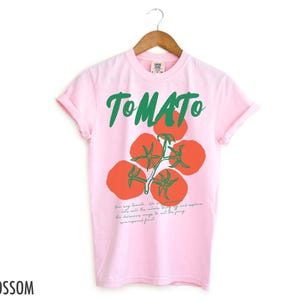 May include: Pink t-shirt with a green and red tomato graphic and the text "TOMATO". The text below the graphic reads "How may tomato. We're not just about salad, eat the whole thing off and explore the delicious ways to eat the juicy, unparelled fruit."