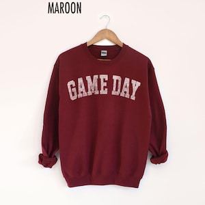 May include: Maroon sweatshirt with the text "GAME DAY" in a distressed, off-white font. The long-sleeved crew neck top is hanging on a wooden hanger against a white background. The word "MAROON" is printed at the top.