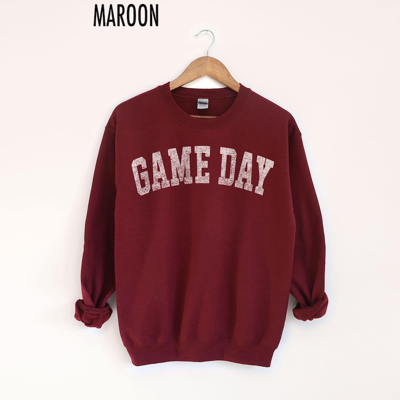 May include: Maroon sweatshirt with the text "GAME DAY" in a distressed, off-white font. The long-sleeved crew neck top is hanging on a wooden hanger against a white background. The word "MAROON" is printed at the top.