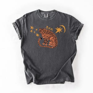 May include: Dark gray t-shirt featuring an orange tiger illustration with stars and a crescent moon. The tiger has black stripes and the word "Raa" above its head. The shirt has rolled-up sleeves and a vintage look.