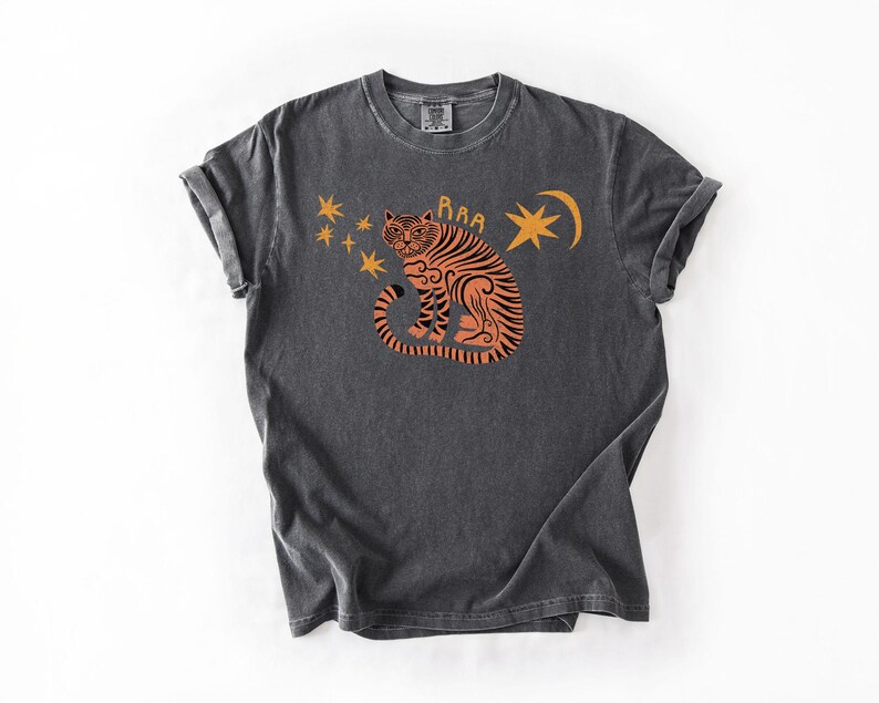 May include: Dark gray t-shirt featuring an orange tiger illustration with stars and a crescent moon. The tiger has black stripes and the word "Raa" above its head. The shirt has rolled-up sleeves and a vintage look.