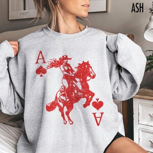 May include: A grey sweatshirt with a red graphic of a woman riding a horse, with playing card symbols and the letter "A". The word "ASH" is printed in the upper right corner.