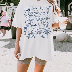 Puede incluir: Camiseta blanca con ilustraciones y texto en azul. El dise&ntilde;o incluye un cangrejo, un traje de ba&ntilde;o, una estrella de mar, gafas de sol, un helado y las palabras "With love from Aruba". La camiseta es holgada.