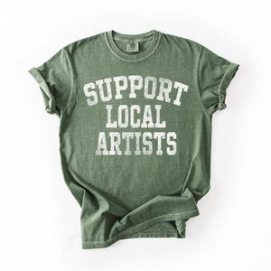 May include: Green t-shirt with the text "SUPPORT LOCAL ARTISTS" in white, block letters. The shirt has a vintage, worn look, with rolled-up sleeves. The shirt is laid flat on a white surface.