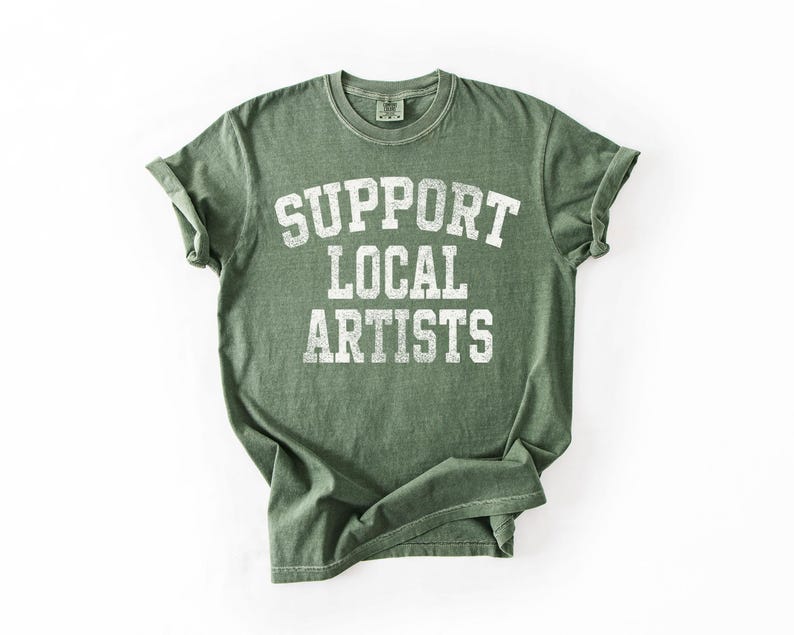 May include: Green t-shirt with the text "SUPPORT LOCAL ARTISTS" in white, block letters. The shirt has a vintage, worn look, with rolled-up sleeves. The shirt is laid flat on a white surface.