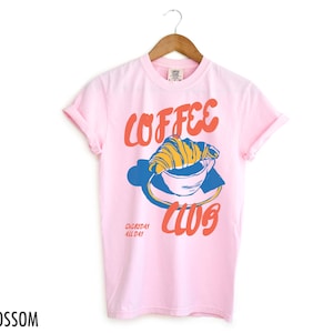 Puede incluir: Camiseta rosa con un gr&aacute;fico de una taza de caf&eacute; con un croissant en la parte superior. El texto "Coffee Club" est&aacute; escrito en rojo y naranja. El texto "Everyday All Day" est&aacute; escrito en negro.