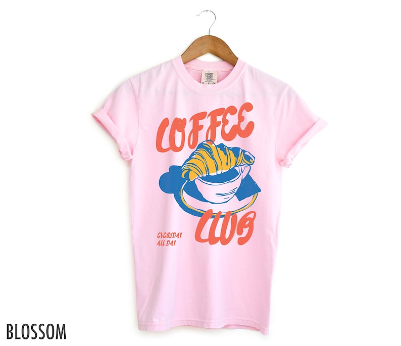 Puede incluir: Camiseta rosa con un gr&aacute;fico de una taza de caf&eacute; con un croissant en la parte superior. El texto "Coffee Club" est&aacute; escrito en rojo y naranja. El texto "Everyday All Day" est&aacute; escrito en negro.