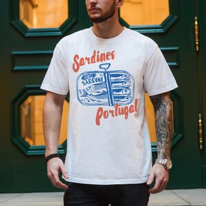 Op de afbeelding: Wit T-shirt met een blauwe en rode afbeelding van een sardineblik met de tekst "Sardines Portugal".