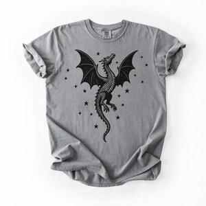Op de afbeelding: Grijs T-shirt met een zwart drakenontwerp. De draak heeft uitgespreide vleugels en is omringd door kleine zwarte sterren. Het shirt heeft een ronde hals en korte mouwen.