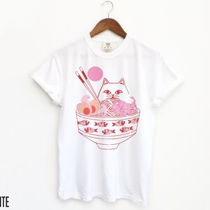 May include: White t-shirt with a pink and red illustration of a cat in a bowl of ramen noodles. The cat is wearing a pink headband and has a pink nose. The bowl is decorated with red fish.