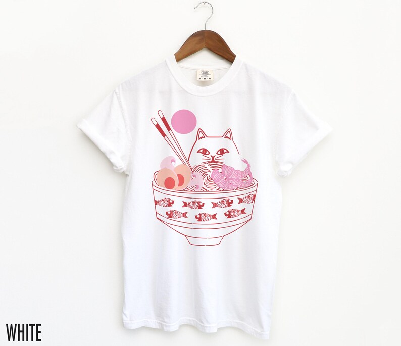 May include: White t-shirt with a pink and red illustration of a cat in a bowl of ramen noodles. The cat is wearing a pink headband and has a pink nose. The bowl is decorated with red fish.