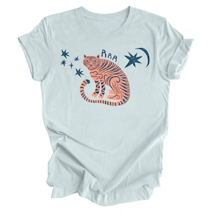 Peut inclure: Un t-shirt bleu clair avec un motif repr&eacute;sentant un tigre orange &agrave; rayures noires assis, la queue enroul&eacute;e autour de son corps. Le tigre est entour&eacute; d'&eacute;toiles bleues et d'un croissant de lune avec une &eacute;toile &agrave; l'int&eacute;rieur. Le texte "RAA" est au-dessus du tigre.