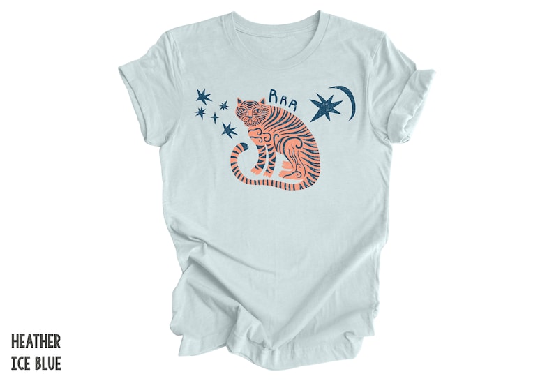 Peut inclure: Un t-shirt bleu clair avec un motif repr&eacute;sentant un tigre orange &agrave; rayures noires assis, la queue enroul&eacute;e autour de son corps. Le tigre est entour&eacute; d'&eacute;toiles bleues et d'un croissant de lune avec une &eacute;toile &agrave; l'int&eacute;rieur. Le texte "RAA" est au-dessus du tigre.