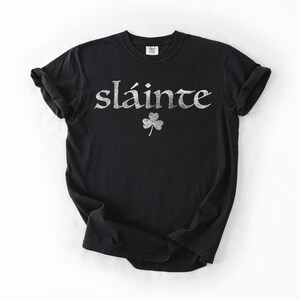 May include: Black t-shirt with the word "sl&aacute;inte" in silver glitter lettering and a glitter shamrock. The shirt is a crew neck and has short sleeves. The shirt is made of a soft material.