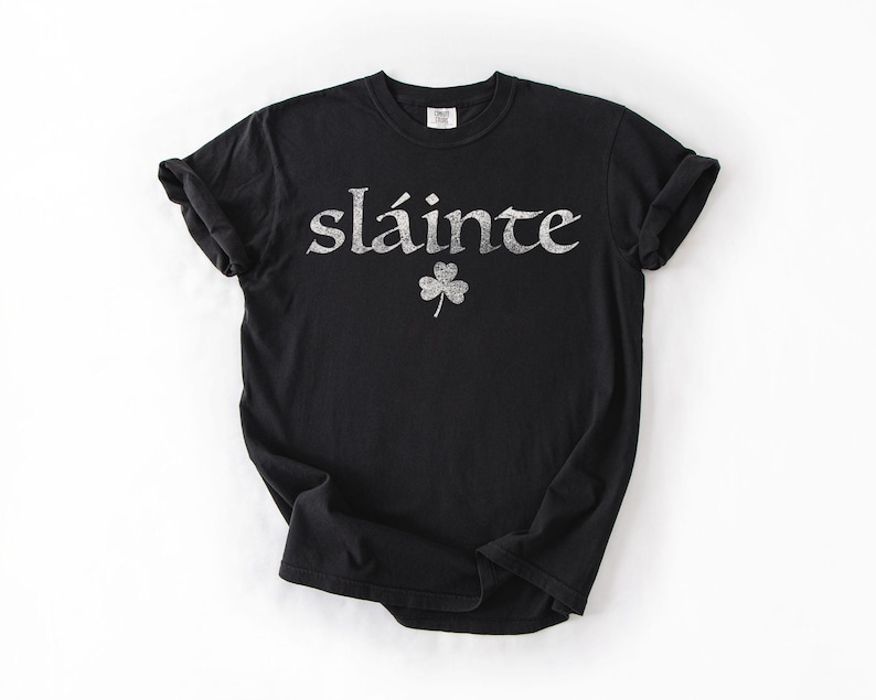 May include: Black t-shirt with the word "sl&aacute;inte" in silver glitter lettering and a glitter shamrock. The shirt is a crew neck and has short sleeves. The shirt is made of a soft material.