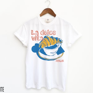 May include: White t-shirt with a graphic of a croissant in a cup of coffee and the text "La dolce vita" and "ITALIA".