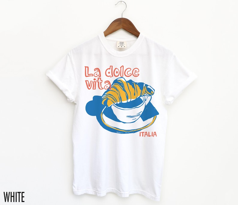 May include: White t-shirt with a graphic of a croissant in a cup of coffee and the text "La dolce vita" and "ITALIA".