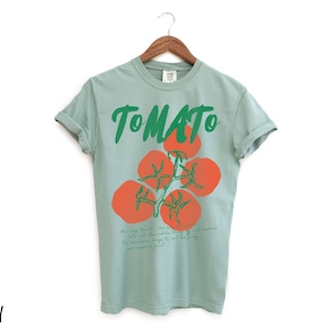 May include: A light blue t-shirt with a green and red tomato graphic and the word "TOMATO" printed in green. The text below the graphic reads "How you say Tomato is a whole other story. But, life was meant to be eaten. If you were meant to just eat the leafy greens, you'd be a rabbit."