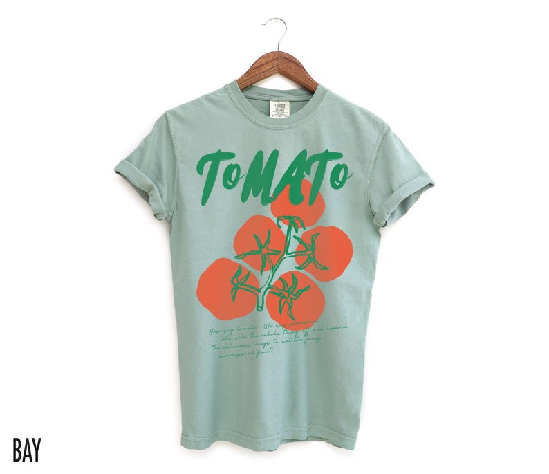 May include: A light blue t-shirt with a green and red tomato graphic and the word "TOMATO" printed in green. The text below the graphic reads "How you say Tomato is a whole other story. But, life was meant to be eaten. If you were meant to just eat the leafy greens, you'd be a rabbit."