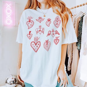 K&ouml;nnte beinhalten: Wei&szlig;es Oversize-T-Shirt mit einem roten Grafikdruck aus mehreren stilisierten Herzen mit unterschiedlichen Designs. Die Herzen haben Details wie Fl&uuml;gel, Augen und florale Elemente.