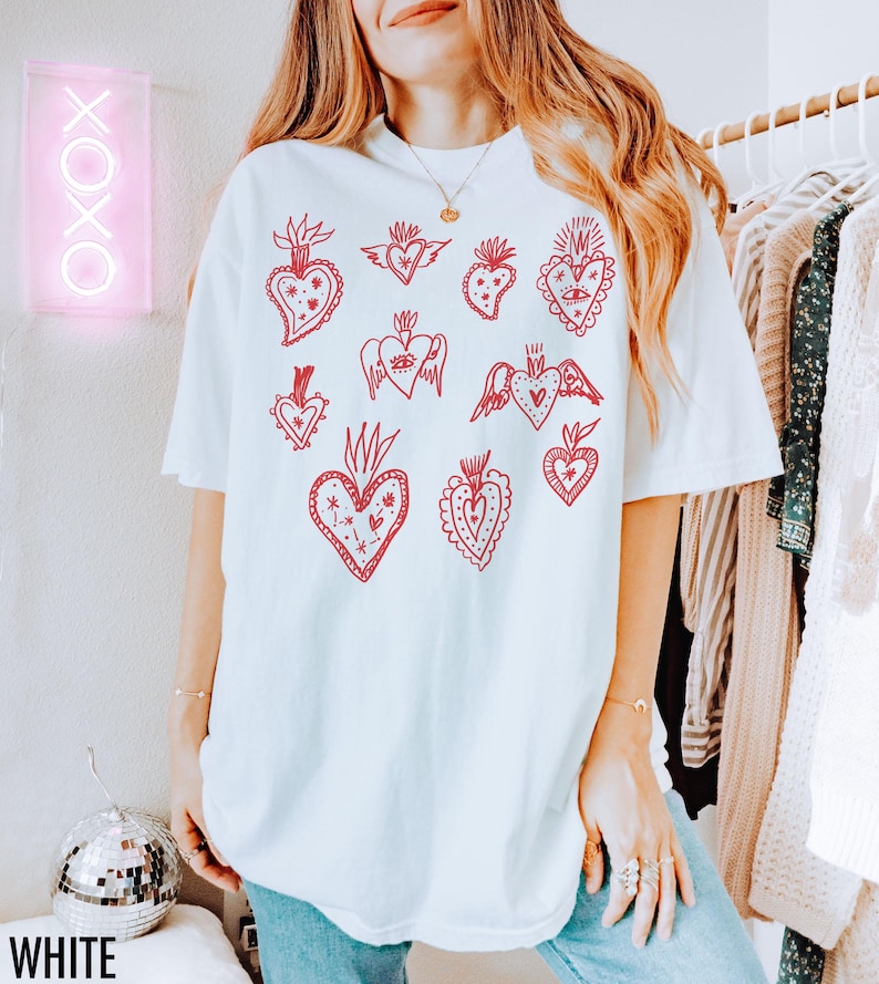 K&ouml;nnte beinhalten: Wei&szlig;es Oversize-T-Shirt mit einem roten Grafikdruck aus mehreren stilisierten Herzen mit unterschiedlichen Designs. Die Herzen haben Details wie Fl&uuml;gel, Augen und florale Elemente.