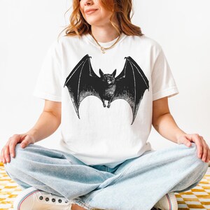 May include: White t-shirt featuring a detailed black illustration of a bat with outstretched wings. The bat design is centered on the shirt, creating a striking contrast against the white fabric. The shirt is a casual style.