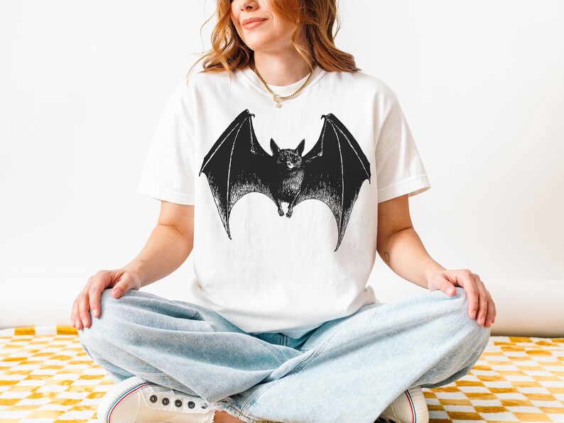 May include: White t-shirt featuring a detailed black illustration of a bat with outstretched wings. The bat design is centered on the shirt, creating a striking contrast against the white fabric. The shirt is a casual style.