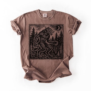 Puede incluir: Camiseta marr&oacute;n con un gr&aacute;fico negro de un hombre lobo en un bosque bajo una luna creciente. El dise&ntilde;o est&aacute; enmarcado dentro de un borde rectangular. La camiseta tiene cuello redondo y mangas cortas.