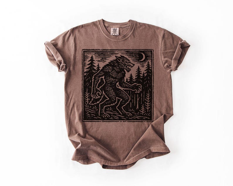 Puede incluir: Camiseta marr&oacute;n con un gr&aacute;fico negro de un hombre lobo en un bosque bajo una luna creciente. El dise&ntilde;o est&aacute; enmarcado dentro de un borde rectangular. La camiseta tiene cuello redondo y mangas cortas.