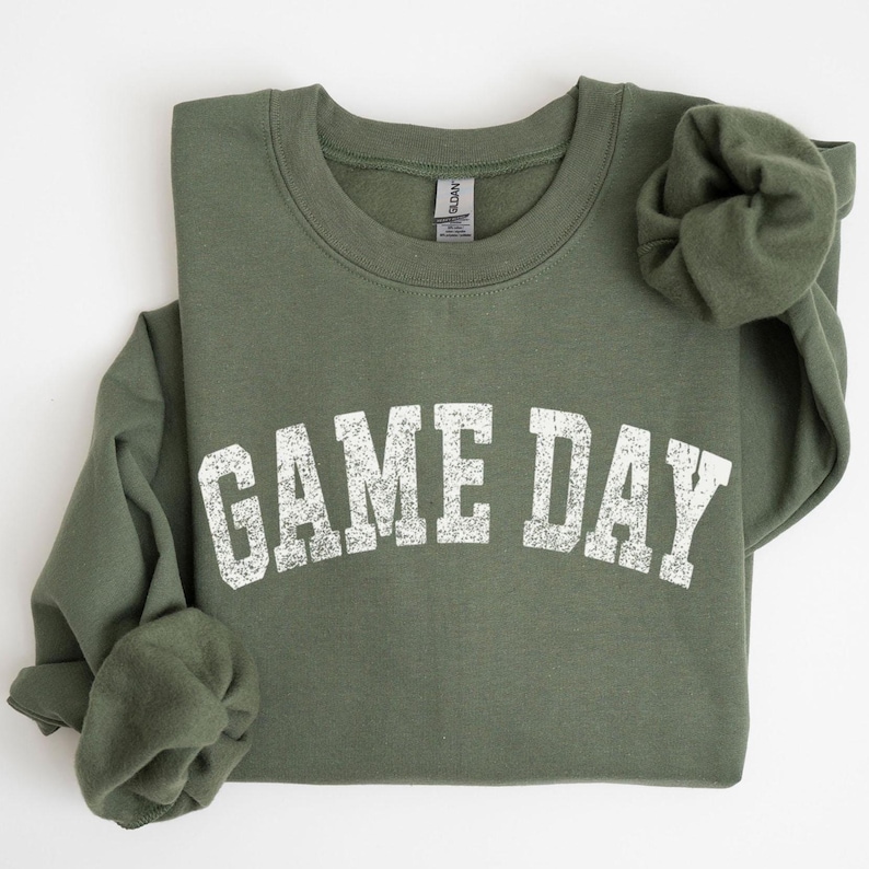 May include: An olive green sweatshirt with the text "GAME DAY" in distressed white lettering. The crew neck sweatshirt is folded, with the sleeves rolled up. The Gildan brand tag is visible on the inside of the collar.