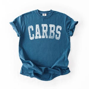 May include: A blue t-shirt with the word "CARBS" printed in distressed white block letters across the chest. The shirt has short sleeves and a crew neck. The fabric appears to be a soft, worn cotton.