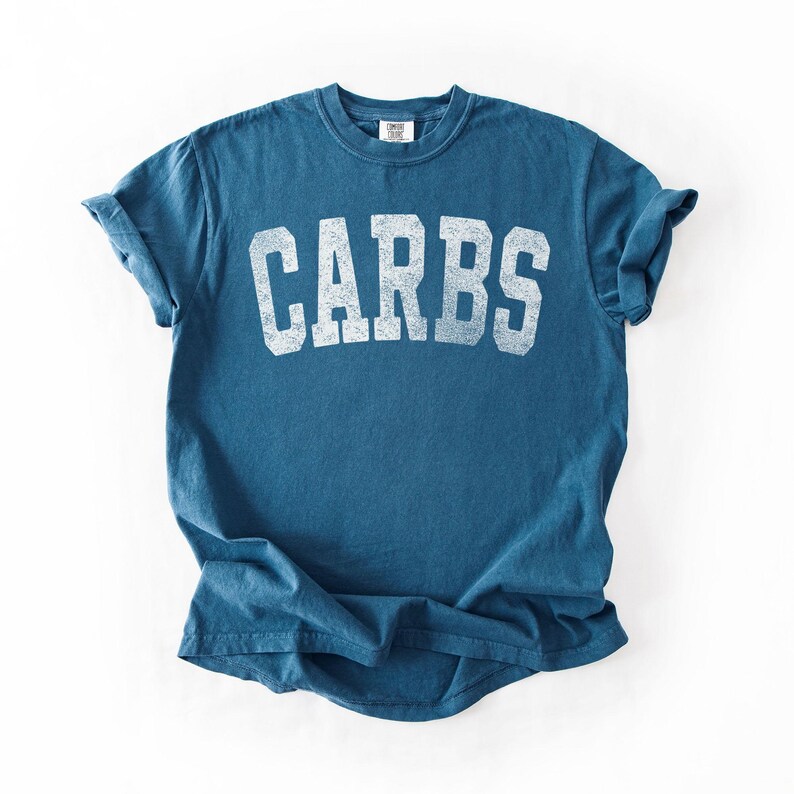 May include: A blue t-shirt with the word "CARBS" printed in distressed white block letters across the chest. The shirt has short sleeves and a crew neck. The fabric appears to be a soft, worn cotton.