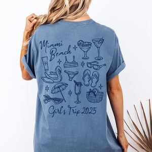 May include: A blue t-shirt with a graphic design of a beach scene with the text "Miami Beach Girls Trip 2025" printed on the back.