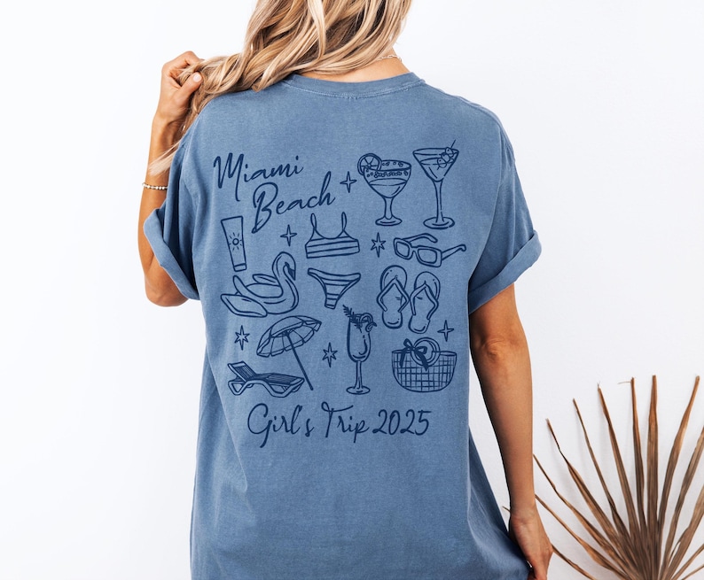 May include: A blue t-shirt with a graphic design of a beach scene with the text "Miami Beach Girls Trip 2025" printed on the back.