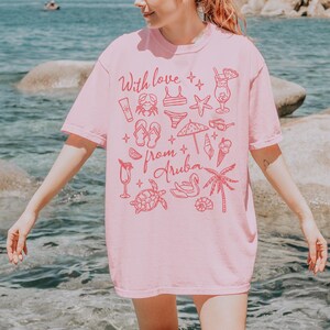 Puede incluir: Una camiseta rosa claro con el texto "With love from Aruba" en rojo. La camiseta presenta dibujos lineales rojos de elementos de playa, como un cangrejo y gafas de sol.