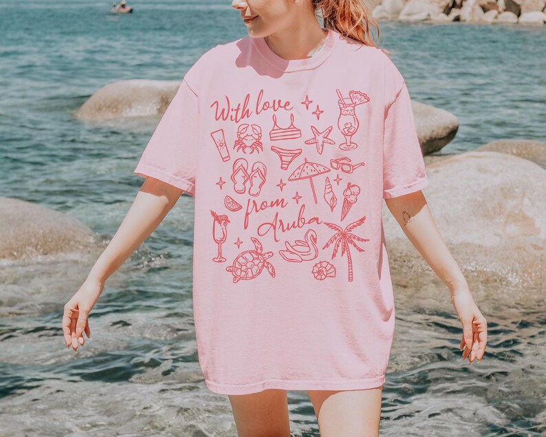 Puede incluir: Una camiseta rosa claro con el texto "With love from Aruba" en rojo. La camiseta presenta dibujos lineales rojos de elementos de playa, como un cangrejo y gafas de sol.