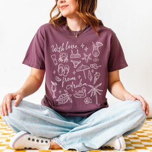 Puede incluir: Una camiseta granate con las palabras "With love from Aruba" e ilustraciones blancas con tem&aacute;tica playera. El dise&ntilde;o incluye un c&oacute;ctel, gafas de sol y una palmera. La persona lleva vaqueros azul claro y zapatillas blancas.