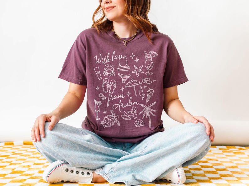 Puede incluir: Una camiseta granate con las palabras "With love from Aruba" e ilustraciones blancas con tem&aacute;tica playera. El dise&ntilde;o incluye un c&oacute;ctel, gafas de sol y una palmera. La persona lleva vaqueros azul claro y zapatillas blancas.