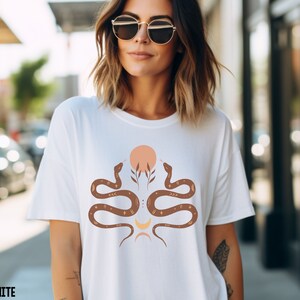 Puede incluir: Camiseta blanca con un gr&aacute;fico de dos serpientes marrones enfrentadas con un dise&ntilde;o de sol y luna en el centro. Las serpientes tienen detalles dorados y el sol es melocot&oacute;n.