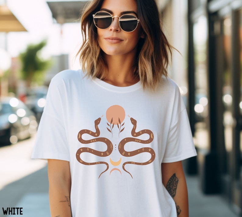Puede incluir: Camiseta blanca con un gr&aacute;fico de dos serpientes marrones enfrentadas con un dise&ntilde;o de sol y luna en el centro. Las serpientes tienen detalles dorados y el sol es melocot&oacute;n.