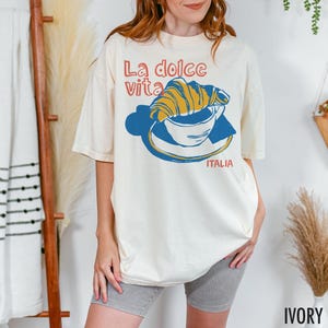 May include: A white t-shirt with a graphic of a croissant in a cup of coffee. The text "La dolce vita" is above the graphic and "ITALIA" is below. The shirt is ivory.