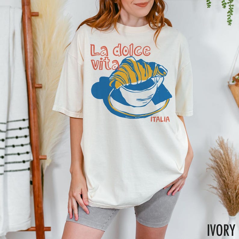 May include: A white t-shirt with a graphic of a croissant in a cup of coffee. The text "La dolce vita" is above the graphic and "ITALIA" is below. The shirt is ivory.