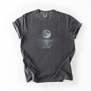 May include: Dark gray t-shirt featuring a celestial design. The graphic depicts a full moon over a body of water, with stars in the sky. The shirt has rolled-up sleeves and a crew neck.