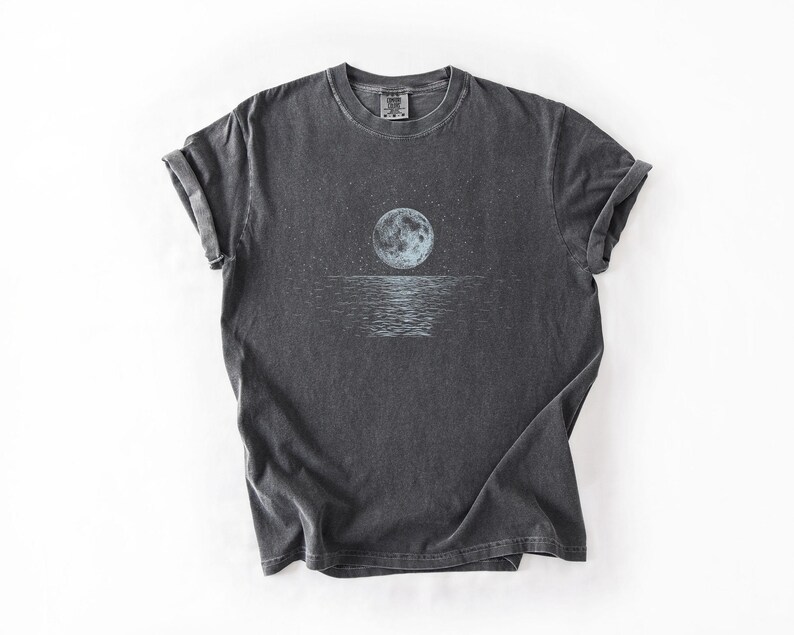 May include: Dark gray t-shirt featuring a celestial design. The graphic depicts a full moon over a body of water, with stars in the sky. The shirt has rolled-up sleeves and a crew neck.